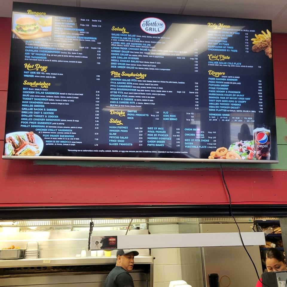 Restaurant menu board displays burgers, sandwiches, salads, fries, and drinks above an open kitchen with two employees.