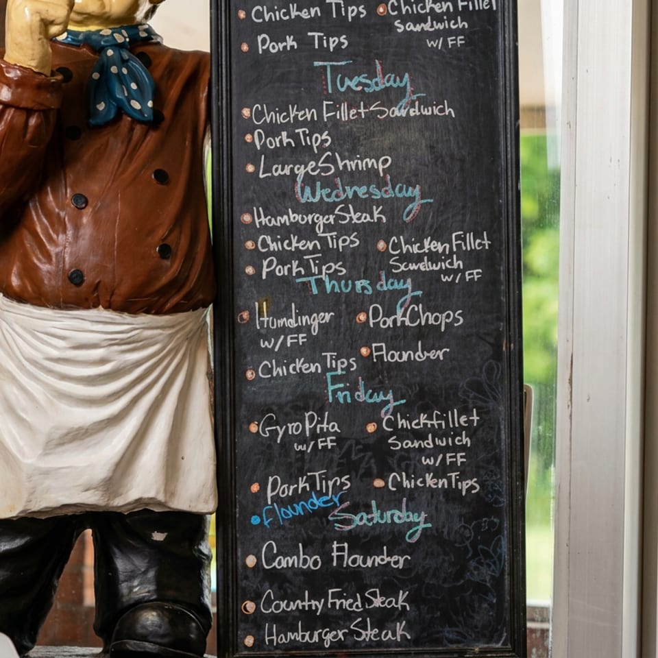 Chalkboard restaurant menu lists daily meat dishes beside a chef statue in a window overlooking green fields.