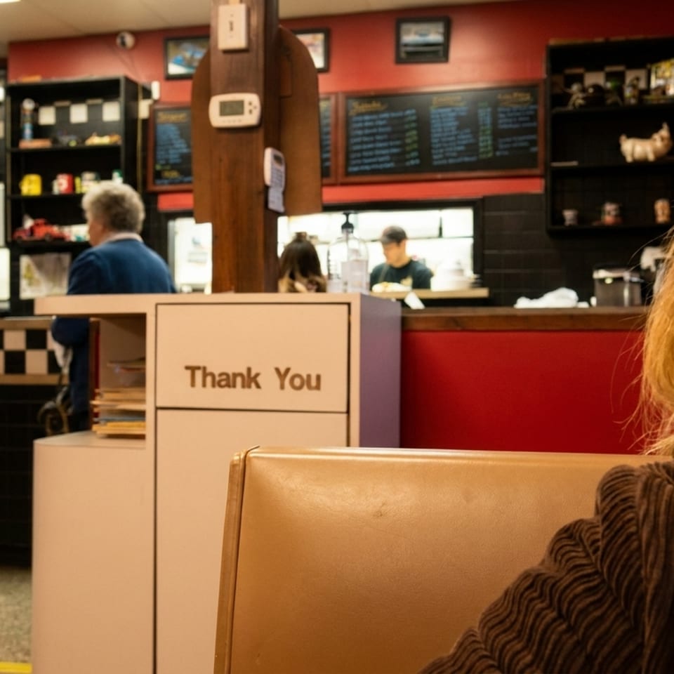 Casual restaurant interior with red and black checkerboard décor, counter staff, chalkboard menus, and a seated customer in the foreground.
